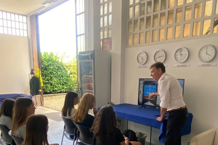 Gruppo di ragazzi e ragazze durante il briefing teorico dell'Aviation Summer Camp 2025 con l'istruttore Stefano Carniti davanti agli schermi e ai world clocks New York · Milano · Sidney
